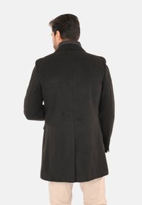 Uomo con cappotto nero e pantaloni beige, in piedi con la schiena rivolta verso un semplice sfondo bianco.