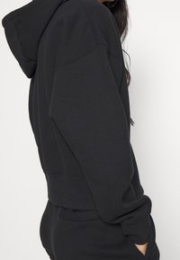 Zwarte cropped hoodie van zachte stof, met een grote kap, afgezakte schouders en een kangaroezak, voorzien van zichtbare stikdetails.
