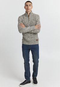 Brede gebreide trui in beige en grijze tinten met een hoge kraag, voorzien van een ritssluiting aan de voorkant en een klein bruin logopatch. Gecombineerd met een donkere jeans en zwarte sneakers.