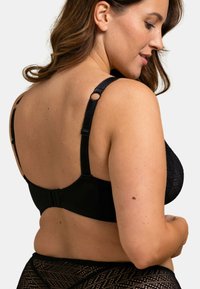 Soutien-gorge noir avec un design en dentelle texturée, des bretelles réglables avec des éléments ronds, et une fermeture à crochets et œillets dans le dos.