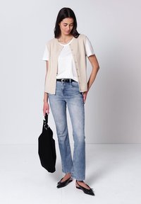 Femme portant un gilet beige sans manches boutonné sur un t-shirt blanc, un jean bleu clair, des chaussures slingback noires, tenant un sac fourre-tout noir.