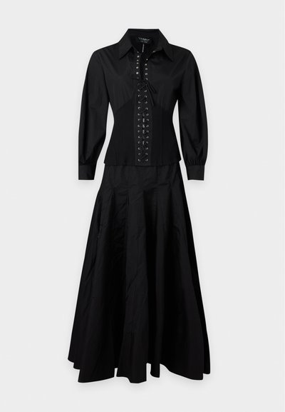 Blouse noire à manches longues avec détail lacé à l'avant, associée à une jupe longue fluide assortie sur un fond uni.