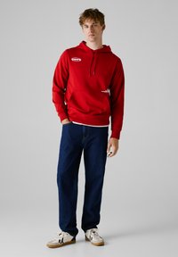 Rote Kapuzenjacke mit vorderer Tasche, weiße Logodetails. Kombiniert mit dunkelblauen geraden Jeans und beigen Sneakers mit schwarzen Akzenten.