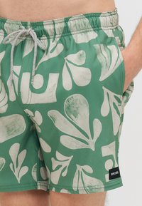 Πράσινα μαγιό κολύμβησης με σχέδιο από φύλλα και σχήματα σε λευκό χρώμα, με ρυθμιζόμενη μέση, πλαϊνές τσέπες και μαύρη ετικέτα "RIP CURL" στο στρίφωμα.