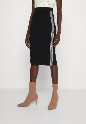 Czarna spódnica do kolan z białym paskiem z logo po boku, noszona z beżowymi botkami na obcasie i białym topem bez rękawów.