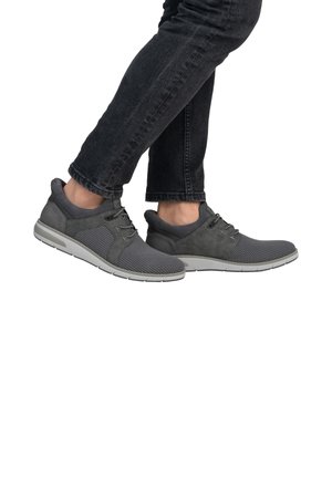 Zapatillas deportivas grises con parte superior de malla y ante, diseño de cordones y suela de goma blanca. Llevadas con jeans oscuros, mostrando el perfil y el movimiento.