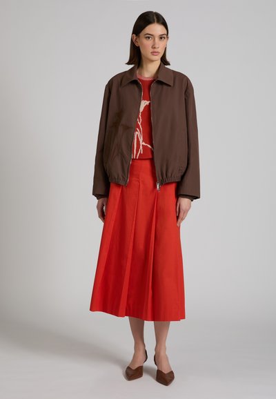 Mujer de pie con chaqueta marrón con cremallera, suéter rojo con patrón, falda midi plisada roja y zapatos planos marrones con punta en un fondo gris.