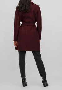 Abrigo de mezcla de lana burdeos con cuello entallado, mangas largas y cinturón. Llevado sobre jeans negros y botas de tacón. Textura suave, diseño minimalista.