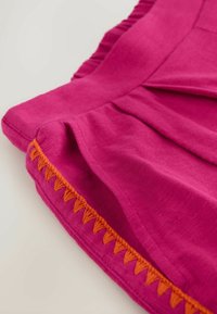 Tissu rose vif avec des volants froncés et une bordure brodée triangulaire orange le long du bord, présenté sur un fond clair.