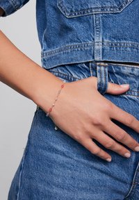 Złoty łańcuszek bransoletki z różowymi, fioletowymi i przezroczystymi koralikami, charakteryzujący się delikatnym wzornictwem i bezpiecznym zapięciem, noszony na ręce na tle materiału dżinsowego.