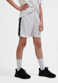 Weiße Sportshorts mit einem schwarzen Chevron-Akzent an der Seite, kombiniert mit schwarzen Sneakers und weißen Sportsocken. Glattes Material, lockere Passform.