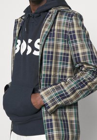 Osoba v námořnickém mikině s bílým nápisem "BOSS" a hnědým, zeleným a námořnickým kostkovaným sakem, s rukama v kapsách mikiny.