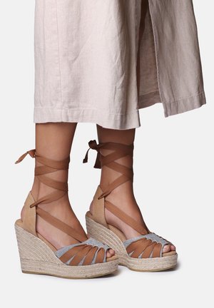 Beine in beigefarbenen und grauen Keilsandalen mit Knöchelriemchen, kombiniert mit einem hellen Rock, der unterhalb der Knie endet.