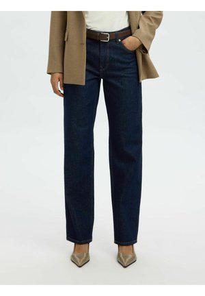 Osoba oblečená v tmavomodrých rifle s rovnými nohavicami, béžových lodičkách so špicatou špičkou, bielom tričku a kamelovom sakom s jednou rukou v vrecku.