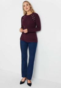 Bordeaux geribbelde trui met oogjesdetails op de schouders, gecombineerd met donkerblauwe flared jeans en zwarte puntige schoenen.