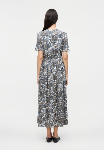 Femme debout de dos, portant une robe midi à fleurs bleues et blanches avec des manches courtes et des ballerines noires sur un fond uni.