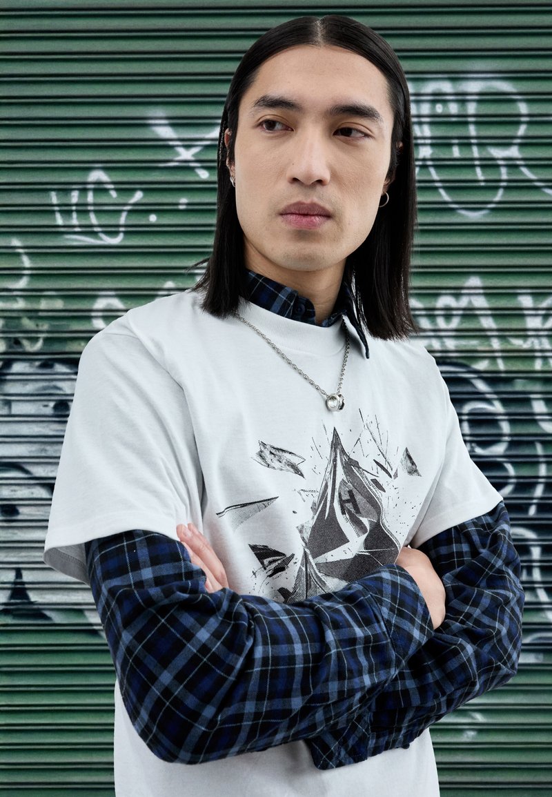 White t-shirt featuring an abstract black graphic, layered over a blue and black plaid long-sleeve shirt, against a green background.