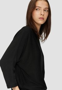 Čierny top s dlhým rukávom a voľným strihom, so širokými rukávmi a textúrovanou tkaninou, prezentovaný na modeli na neutrálnom pozadí.
