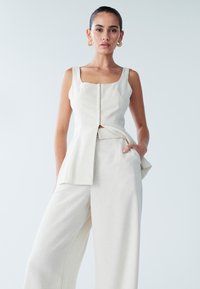 Hellbeiges Oberteil aus Leinenmischung mit quadratischem Ausschnitt, figurbetontem Oberteil und asymmetrischem Vorderteil. Kombiniert mit passenden weit geschnittenen Hosen.