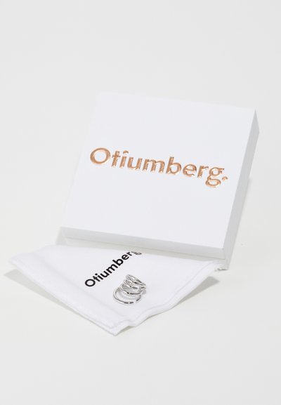 Silberringe auf einem weißen Tuch mit einem schwarzen Logo, neben einer weißen Box mit erhabenem roségoldenen Schriftzug ausgestellt.
