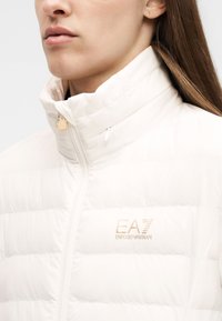 Κοντινό πλάνο γυναίκας που φοράει λευκό μπουφάν puffer με ψηλό γιακά και λογότυπο EA7 Emporio Armani στο στήθος.