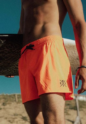 Bovenlichaam ontbloot persoon die een surfplank vasthoudt, draagt feloranje zwembroek met zwart trekkoord onder een helderblauwe lucht.