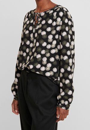 Person iført en sort bluse med lange ærmer med hvide og grønne blomsterprint, kombineret med sorte højttaljede bukser.