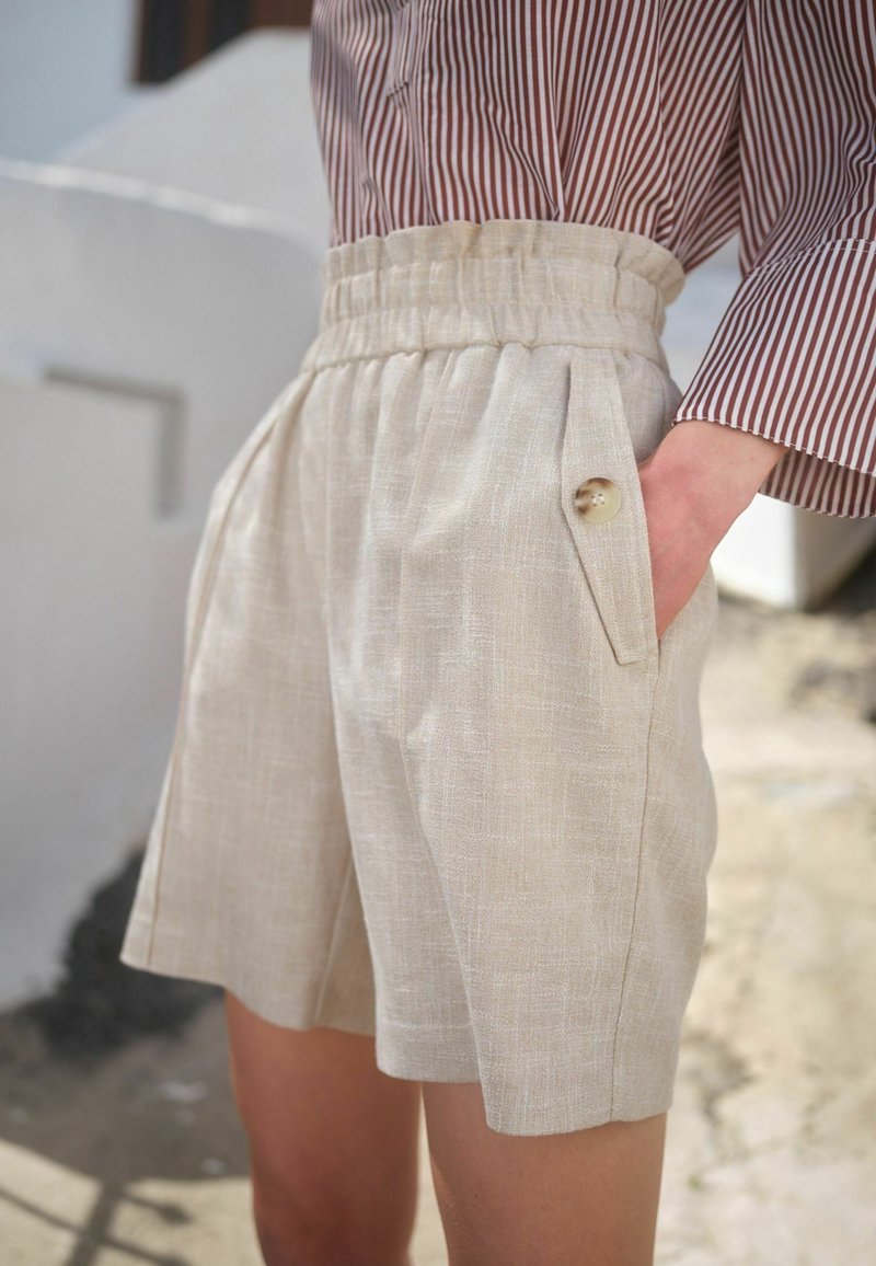 Shorts en lin beige avec un design plissé, une taille élastique et un grand accent en bouton sur la poche. Texture lisse avec une apparence légère.