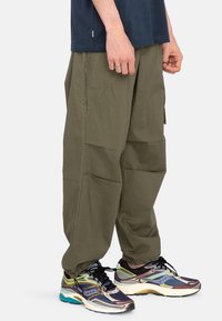 Khaki cargobroek van lichtgewicht stof met meerdere zakken, een ontspannen pasvorm en een gladde textuur, gecombineerd met kleurrijke sportschoenen.