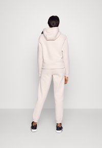 Ljust beige hoodie och sweatpants-set, tillverkat av mjukt material. Har en huva, ribbade ärmslut och elastiskt midjeband. Bärs med svarta sneakers.