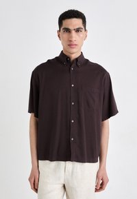 Camicia a maniche corte con bottoni di colore marrone scuro, realizzata in tessuto leggero. Presenta una tasca sul petto e un colletto standard con dettagli in bottone.