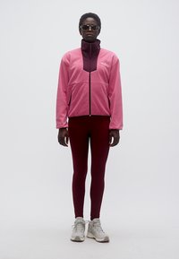 Μαύρο άτομο που στέκεται, φορά γυαλιά ηλίου, μπουφάν φερμουάρ δίχρωμο σε ροζ τόνους, σκουρόχρωμο κόκκινο κολάν και ανοιχτόχρωμα αθλητικά παπούτσια.