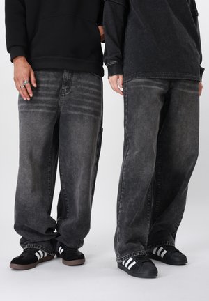 Deux personnes portant des jeans noirs délavés et larges, des baskets Adidas noires et blanches, ainsi que des hauts noirs à manches longues, se tiennent côte à côte.