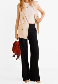 Blazer sans manches beige à double boutonnage sur un haut blanc à côtes, associé à un pantalon noir à jambes larges. Tenant un sac à main frangé de couleur rouille.