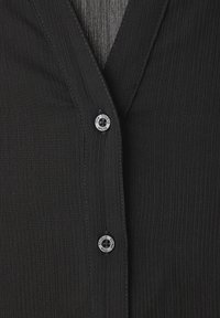 Camisa negra texturizada con diseño de cuello en V, ribetes verticales y detalle de botón en contraste con la marca "Studio".