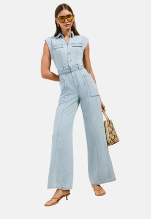 Kvinde iført lyseblå denim jumpsuit uden ærmer med bælte, gultone solbriller, holder en taske med slangeskinds mønster og brune hælede sandaler.