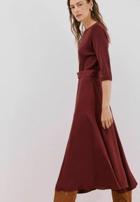 Bordeauxfarbenes Kleid mit halben Ärmeln und tailliertem Bund. Ausgestellter Rock reicht bis zur Mitte der Wade. Glatte Stofftextur mit minimalistischem Design. Braune Stiefel.