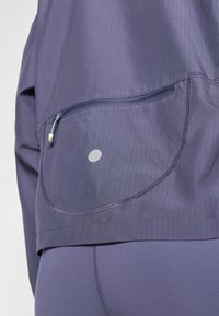 Paarse sporttop met een klein ritsvakje en een stippenpatroon. Heeft een geribbelde textuur en een gladde stof, ontworpen voor comfort en functionaliteit.