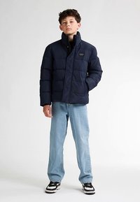 Giubbotto imbottito navy con collo alto e design trapuntato, abbinato a jeans azzurri e sneakers nere. Dettagli di branding minimi visibili.