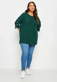 Übergroßer dunkelgrüner Pullover mit Dreiviertelärmeln und Knopfdetails, kombiniert mit hellblauen Skinny-Jeans und weißen Sneakers.