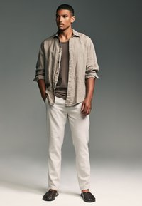 Camicia in lino marrone chiaro con bottoni, canotta marrone scuro, pantaloni color crema e sandali intrecciati scuri, in piedi contro uno sfondo grigio neutro.