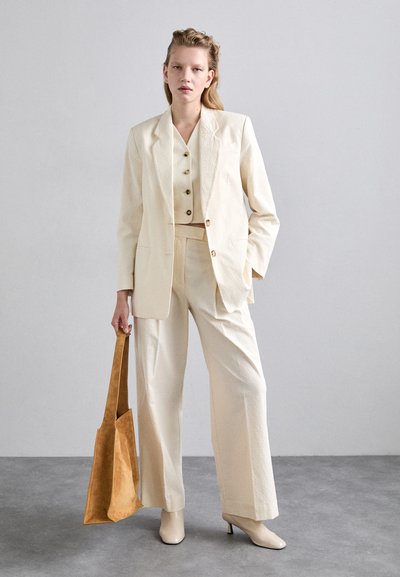 Crème textuur pak met een aansluitende blazer, een knooploze crop top en brede broekspijpen. Gecombineerd met een brown suède tote en beige hakken.