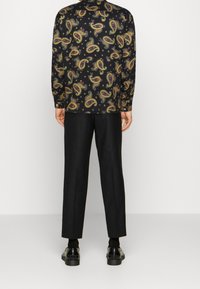 Camisa de manga larga negra con un patrón de paisley en multicolores, combinada con pantalones negros a medida y zapatos negros. Textura de tela suave.