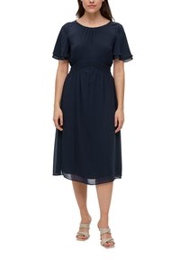 Navyblauwe jurk met korte, fladderende mouwen, gerimpelde taille, vloeiende stof, halflange lengte en doorschijnende zoom. Gekleed met lichtgekleurde sandalen.