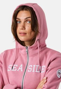 Rosa Zip-Hoodie aus weichem Stoff mit Kapuze, mit dem Schriftzug "SEASIDE" in weißer Schrift und einem Patch am linken Ärmel.