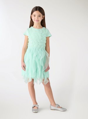 Ragazza giovane in piedi, indossa un vestito verde menta con corpetto floreale e gonna a balze, ballerine argentate glitterate con fiocchi su sfondo bianco.