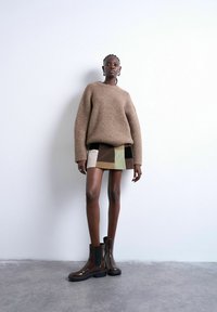 Femme debout contre un mur blanc, portant un pull marron surdimensionné, une mini-jupe patchwork et des bottines marron foncé.