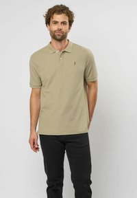 Vaaleanbeige polo-paitaa, jossa on kaulus, kaksi nappia ja pieni logo rinnassa, yhdistettynä tummiin mustiin farkkuihin ja yksinkertaiseen taustaan.