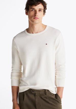 T-shirt blanc à manches longues avec des poignets et un col côtelés. Présente un petit logo sur la poitrine. Porté avec un pantalon vert olive.