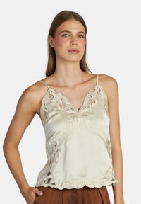 Mujer vestida con un camisón de satén crema con detalles de encaje bordado y tirantes finos, combinado con pantalones marrones frente a un fondo liso.